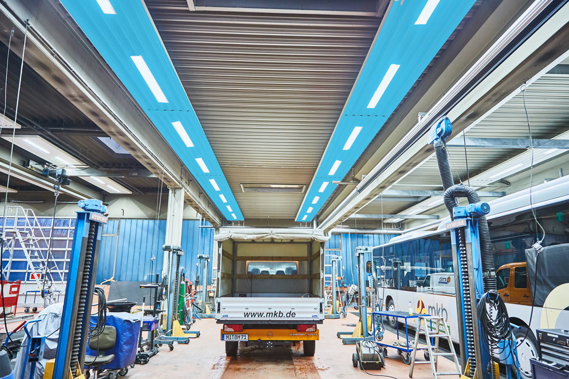 Blau eingefärbte Deckenstrahlplatten in einer Werkhalle, die Kühlung symbolisieren, mit Fahrzeugen und Werkzeug im Hintergrund.